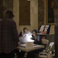 Two poll workers sitting at a table shine a flashlight on a ballot