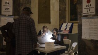 Two poll workers sitting at a table shine a flashlight on a ballot