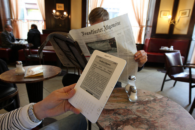 Man reading a newspaper next to man reading news on a Kindle